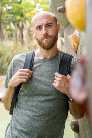 Portrait of a bearded man with shaved head standing next to an outdoor climbing wall.