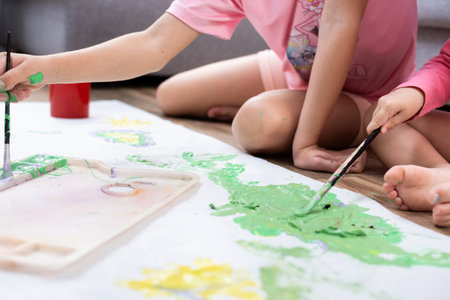 Two sisters sitting on the living room floor, painting together on a large sheet of paper with...
