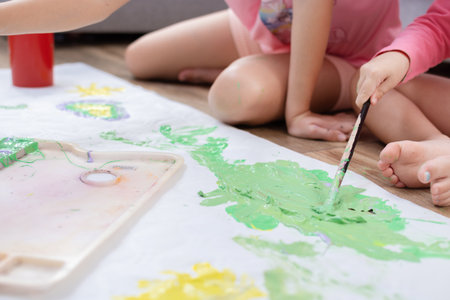 Two sisters sitting on the living room floor, painting together on a large sheet of paper with...