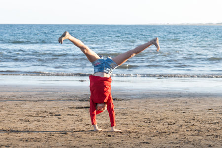 Young girl wearing a red shirt and Santa Claus hat doing a cartwheel on the sandy beach, enjoying a fun and active holiday by the seaの写真素材