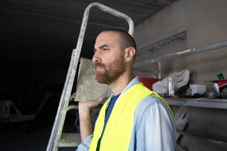 A focused construction worker wearing a reflective safety vest while carrying a ladder inside a...