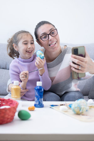 Mother and young daughter taking a selfie with a smartphone while decorating colorful Easter eggs at...