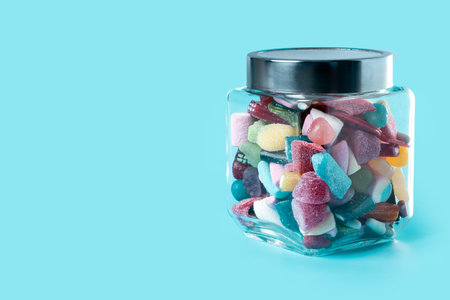 Glass jar filled with assorted colorful gummy candies and sweets on a clean blue background. Bright, vibrant composition with copy space, ideal for concepts like sugar, snacks, childhood, and indulgence.