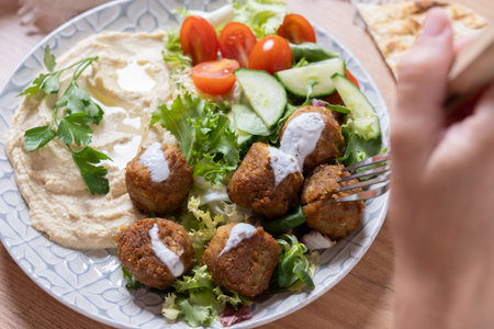 Close-up of hands holding a Mediterranean meal with crispy falafel, creamy hummus, fresh mixed salad, yogurt sauce, and pita bread. Healthy vegetarian cuisine served on a wooden table.