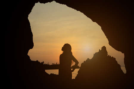 Silhouette of a young woman standing in front of a cave on the beach at sunsetの写真素材