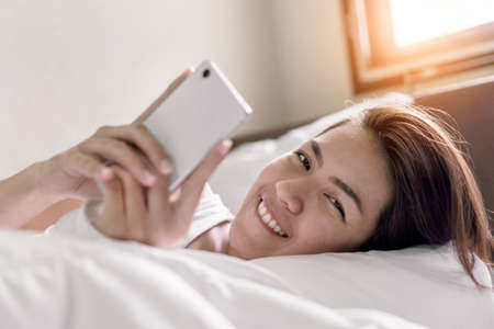 Smile young asian woman playing with her smathphone on white pillow on bed in bedroom on morning time after wake up, Selective focus.の写真素材