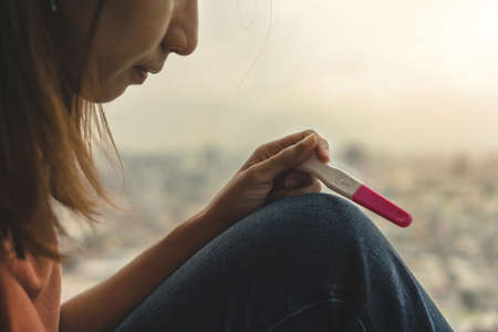 Unhappy young asian woman holding pregnancy test showing a negative result and sit down near big window city view on evening time, Wellness and healthy concept, Infertility problem, Selective focus.の写真素材