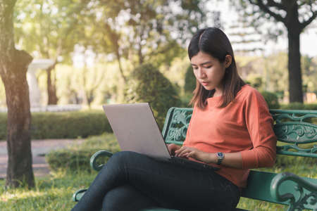 Happy young asian woman working with her laptop on a bench in the park outdoors on vacation time.の写真素材