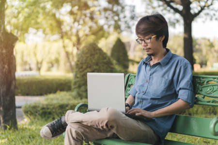 Young asian man working with his laptop on a bench in the park outdoors on vacation time.の写真素材