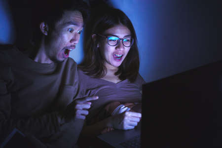 Young asian relaxed woman and man smiling happy on they bed using laptop computer to watching movie or internet social network with low light environment.Selective focus.の写真素材
