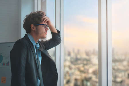 Depressed caucasian man losing job and heartbroken at same time sitting alone near window in dark at evening time with low light environment, PTSD Mental health and depression concept.の写真素材