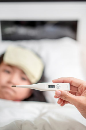 Asian mother measuring temperature girl with digital thermometer in her mouth on bed at morning...