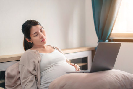 tired asian pregnant woman  and using laptop on bed for working at her bedroom, remote working, Online shopping, Women pregnancy concept, New normal and work from home concept.の写真素材