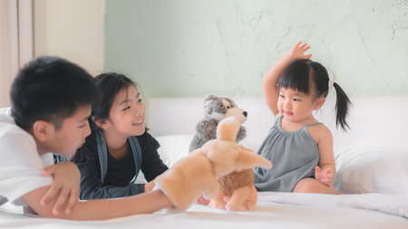 lder asian sister and Elder asian brother playing animal hand puppet doll toys with younger asian sister, sitting on bed, Educational preschool games, Having fun with kid at home concept.の写真素材
