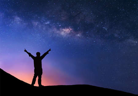 Man is standing next to the Milky Way galaxy with his hands raised to the air.