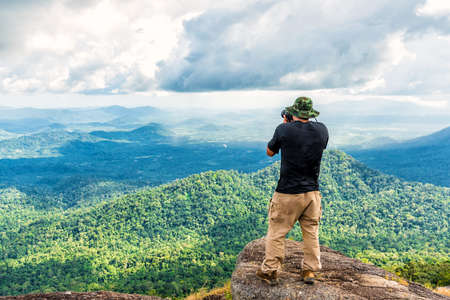 Male photographer enjoying and taking top view valley rain forest photo from mountain cliff.の写真素材