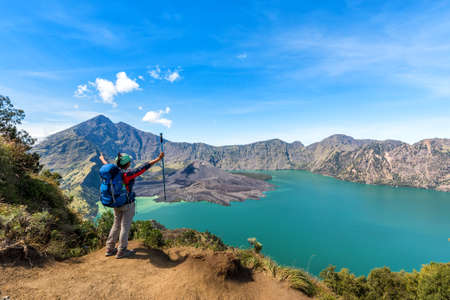 Hiker woman with blue backpack and trekking pole standing, enjoy and happy with active volcano Baru Jari, Lake Segara Anak and summit of Rinjani mountain view after finished climbing at Rinjani mountain, Lombok, Indonesia.の写真素材