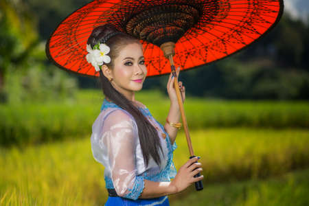 Beautiful asian girl in Myanmar traditional costume, with Red bamboo umbrella on on Rice fields Sky background, identity culture of Thailandの写真素材