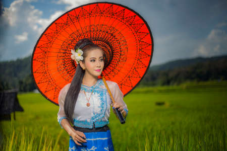 Beautiful asian girl in Myanmar traditional costume, with Red bamboo umbrella on on Rice fields Sky background, identity culture of Thailandの写真素材