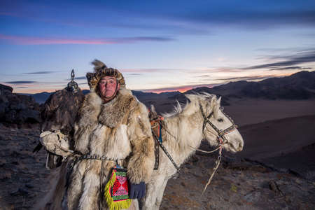 Mongolian Eagle hunter in traditional clothing, holding a golden eagle on his arm and Rider horse on...