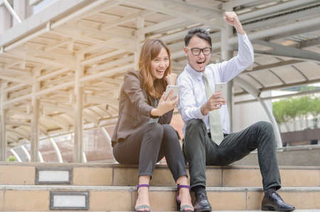 Portrait of an Asian man and woman sitting together and look at their smart phone with smile,buliding background.success concept.の写真素材