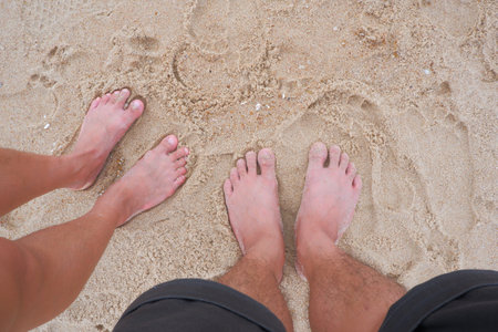 Feet of a man and woman couple standing on a sandy beach with shells in the sand.