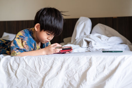 Asian boy lying on bed happily playing with mobile phone enjoying games and digital contentの写真素材