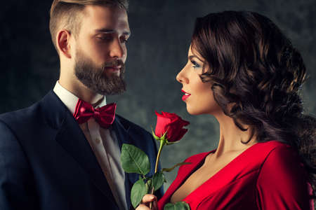 Young elegant couple in evening dress portrait. Woman in red holding rose and looking on man. Focus...