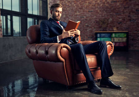 Young handsome businessman with beard in black suit sitting on chair reading book. Focus on face.の写真素材