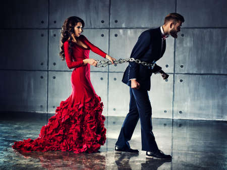 Young woman in red dress holding man on heavy chain. He tries to escape. Elegant evening clothing.の写真素材