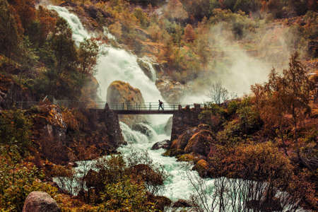 Big waterfall in Norway and man walking on bridge. Autumn red colorsの写真素材