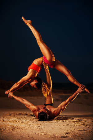 Young sports couple doing acroyoga exercises on sand beach at twilightの写真素材
