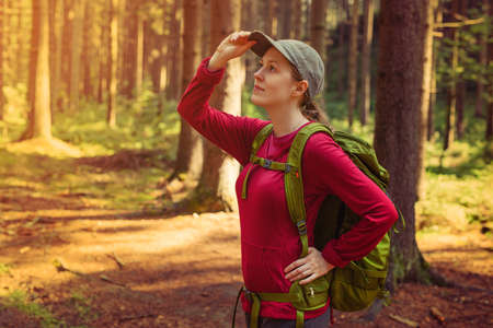 Young woman tourist in red clothing with green backpack looking aside in forest