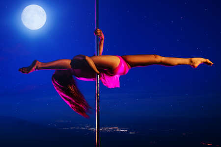 Young slim woman pole dancing at night on sky and hills background