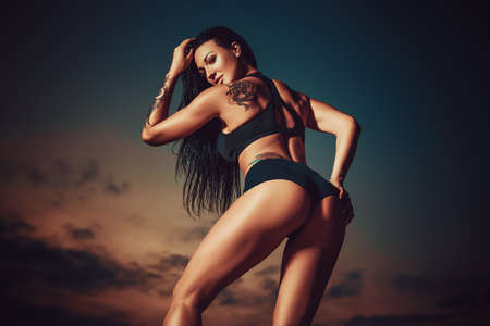 Young sexy slim sports woman posing outdoors on sky background at twilight. Tattoo on body.の写真素材