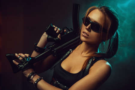 Young strong cool woman with big gun and sunglasses in dark dramatic urban interior