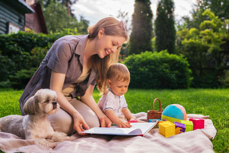 Young mother with one year child and dog sitting in park and reading book