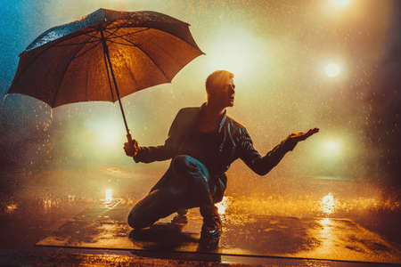 Young strange man with umbrella under the rain. Vibrant lights on background.
