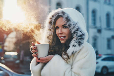 Young woman with warm mug winter portrait in city at sunset lightの写真素材