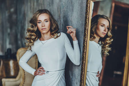 Young slim woman in white dress standing at mirror. Vintage film style colors.