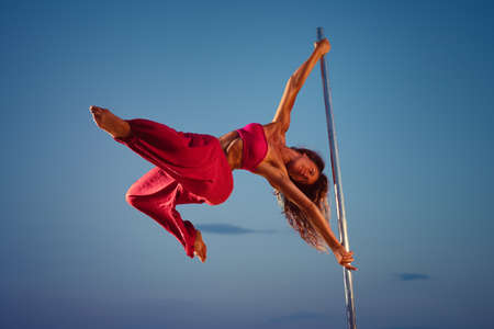 Young sports woman in pink clothing pole dancing at twilight on sunset blue sky background