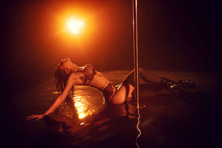 Young sexy slim woman pole dancing in dark interior with red lights and wet floor