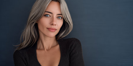 A woman with long blonde hair and bright blue eyes poses confidently in a modern indoor space. Her relaxed expression and elegant outfit stand out against the dark backdrop.の素材