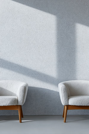 Two stylish white chairs with wooden legs are positioned on a smooth floor beside a textured wall. Soft afternoon light creates gentle shadows, enhancing the serene atmosphere.の素材