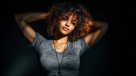 A woman with curly hair poses confidently in a dark setting, showcasing her expressive features and relaxed demeanor. The soft light highlights her facial structure.の素材