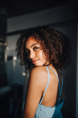 A young woman stands in a well-lit space, showcasing her curly hair and a light blue dress. Soft shadows play across her face as she smiles gently, exuding confidence and warmth.の素材