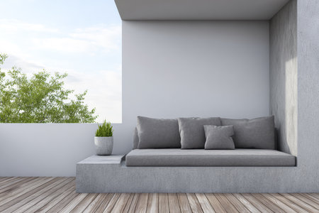A stylish seating area features a smooth concrete couch with plush gray cushions. It is situated on a wooden deck, offering a peaceful view of lush green trees under a clear sky.の素材