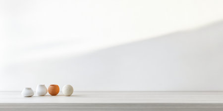 Small ceramic vases, including an orange one, are arranged on a light wooden table. The background is a plain white wall, creating a calm and minimalistic atmosphere.の素材