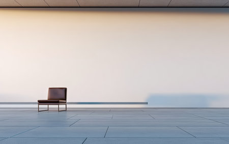 A solitary brown chair sits in an empty room with smooth gray flooring. Soft golden light from the setting sun casts gentle shadows on a plain white wall, creating a calm and serene atmosphere.の素材