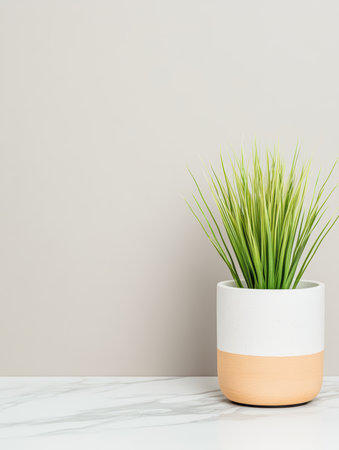 A fresh green plant sits in a white pot with a peach band, displayed on a marble table. The neutral beige wall creates a calm, modern atmosphere.の素材